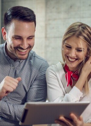 A man and woman happily looks at a tablet, both wearing bright smiles on their faces, radiating joy and excitement.